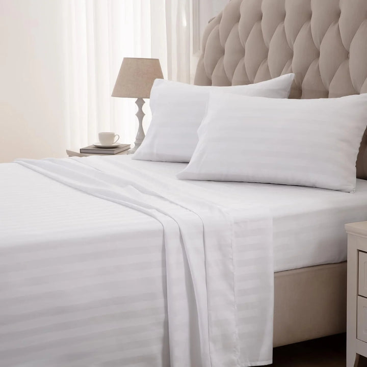 White bed sheets and pillows on a neatly made bed with a lamp and cup on a nightstand.